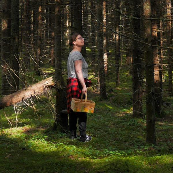 Nainen t-paidassa ja sienikori kädessä hämyisessä metsässä.